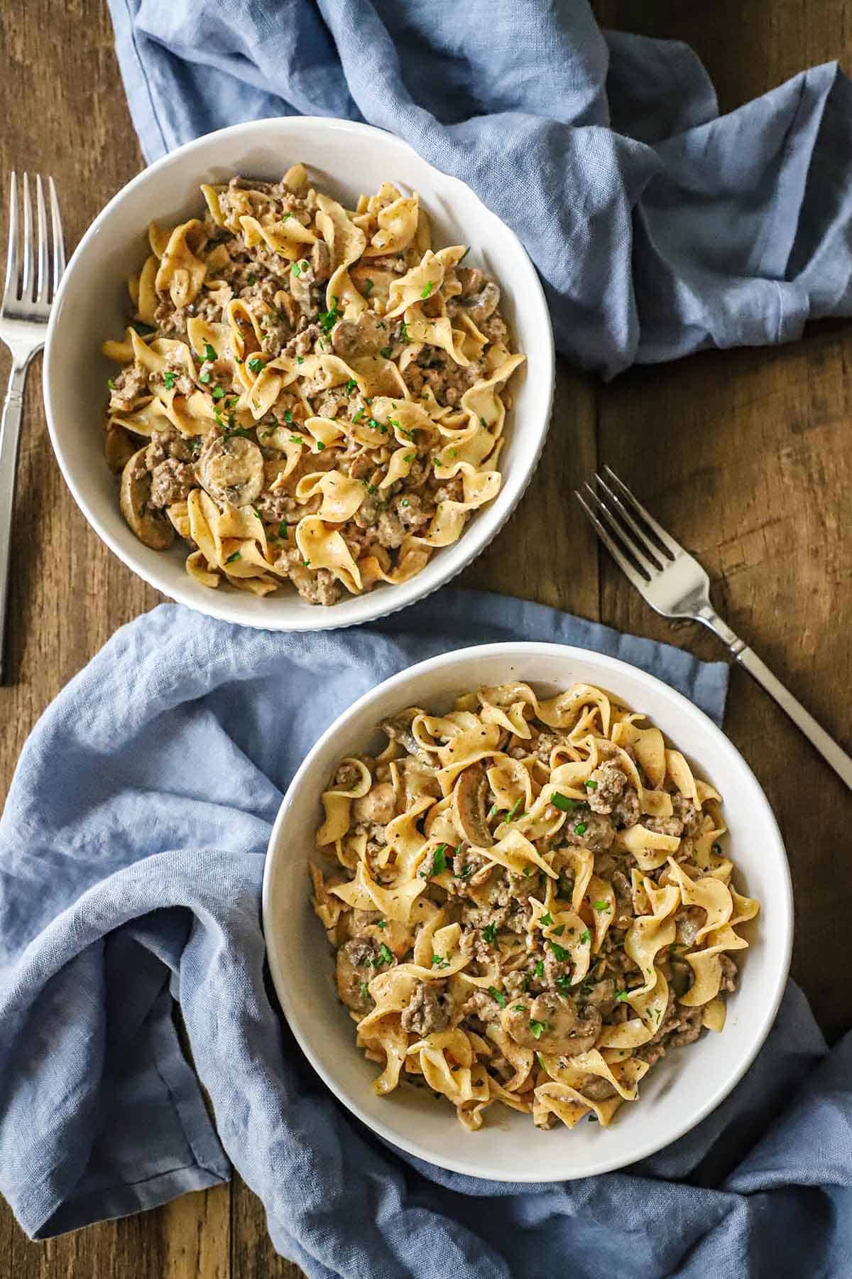 An overhead view of two white pasta bowls filled with servings of Weeknight Beef Stroganoff each with a fork resting nearby.