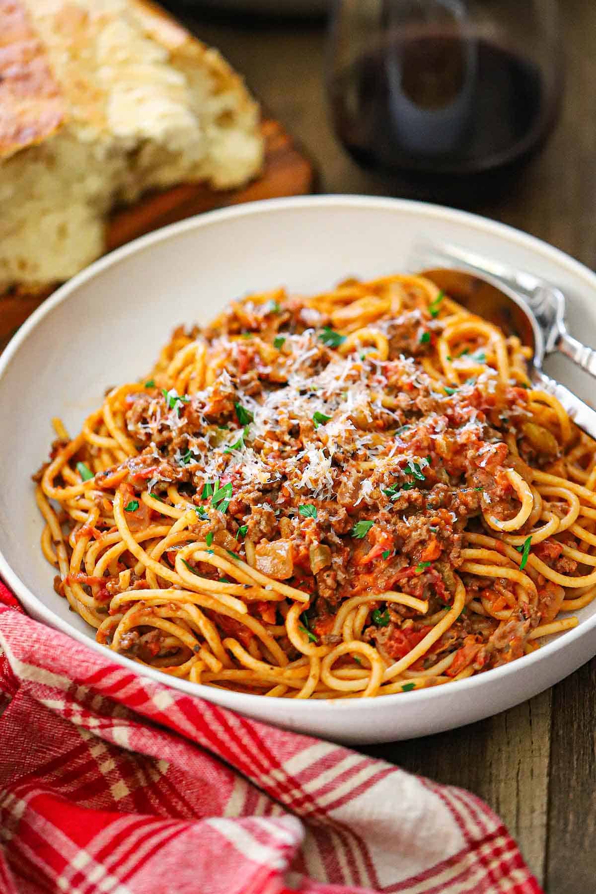 A straight-on view of a shallow white pasta bowl filled with a serving of pasta bolognese next to a stemless glass of red wine.
