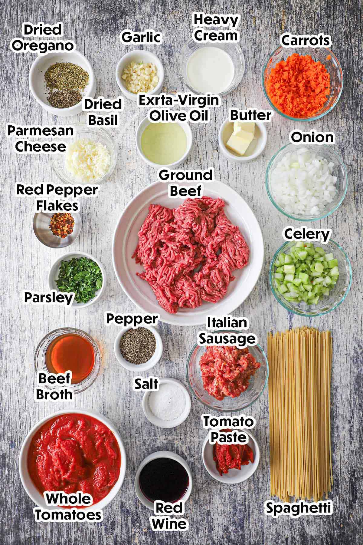 An arrangement of ingredients for weeknight spaghetti bolognese on a grey wooden background including ground beef, sausage, carrots, celery, onion, garlic, beef broth, tomatoes, tomato paste, red wine, heavy cream, Parmesan cheese, spaghetti, herbs, and spices.