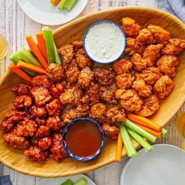 An overhead view of a large oval wooden platter filled with three types crispy boneless chicken wings and a bowl of blue cheese dressing and another bowl filled with barbecue sauce.
