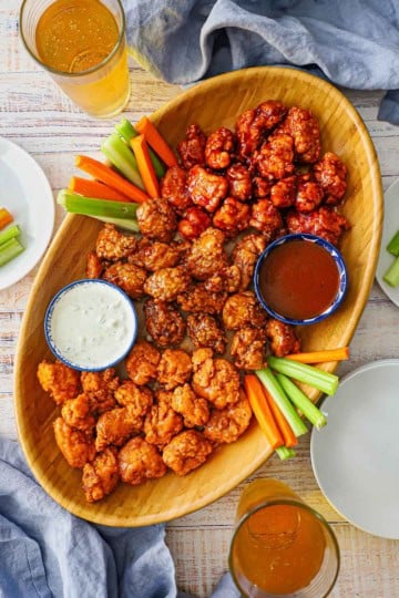 An overhead view of a large oval wooden platter filled with three types crispy boneless chicken wings and a bowl of blue cheese dressing and another bowl filled with barbecue sauce and two glasses of beer sitting nearby.