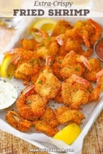 An overhead view of Fried Butterfly Shrimp on a metal pan lined with white wax paper with a small bowl of cocktail sauce in one corner and and a small bowl of tartar sauce in the other corner.