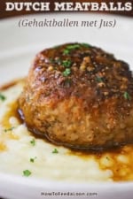 A close-up view of a large Dutch meatball resting on a bed of mashed potatoes with a thin brown gravy drizzled over the top all in a white shallow bowl.