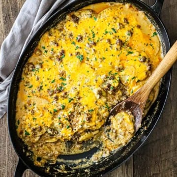 An overhead view of a large oval casserole dish filled with a cheesy potato hamburger casserole with a portion of it missing and a wooden spoon resting in the pan.