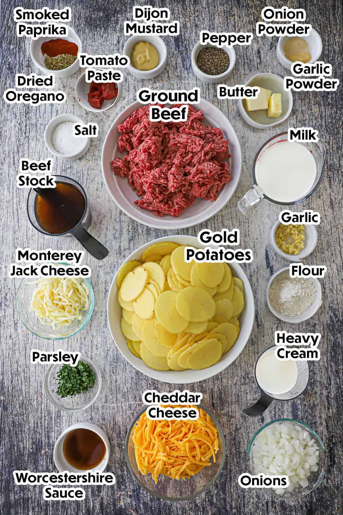 An arrangement of ingredients for hamburger potato casserole on a grey wooden background including ground beef, sliced potatoes, beef, stock, milk, garlic, flour, cream, cheese, onions, butter, tomato paste, and seasonings.