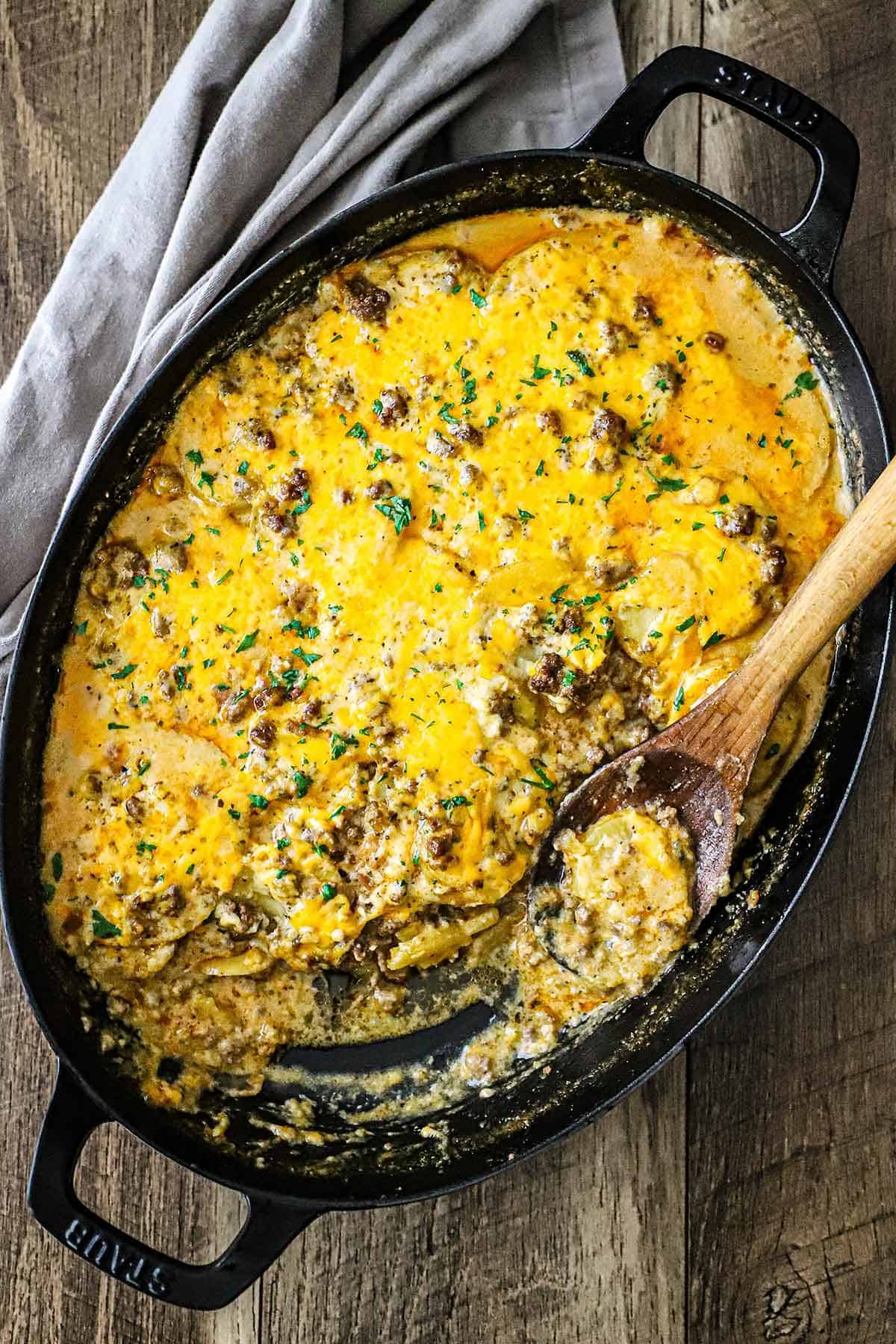 An overhead view of a large oval casserole dish filled with a cheesy potato hamburger casserole with a portion of it missing and a wooden spoon resting in the pan and a grey cloth napkin nearby.