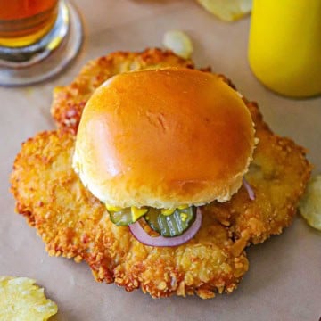 An overhead view of a breaded pork tenderloin sandwich topped with red onion, pickle slices, and yellow mustard.