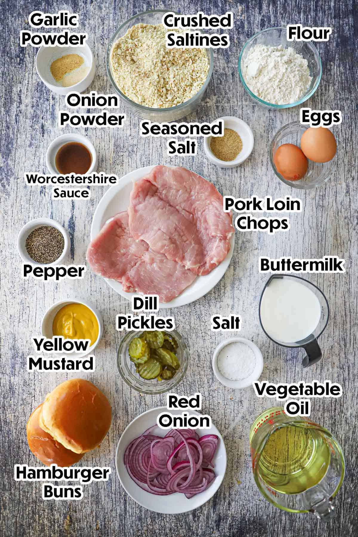 An arrangement of ingredients for breaded pork tenderloin sandwich on a grey-wooden background included thinly pounded pork loin chops, buttermilk, eggs, crushed saltines, flour, oil, mustard, buns, onions, pickles, and seasonings.