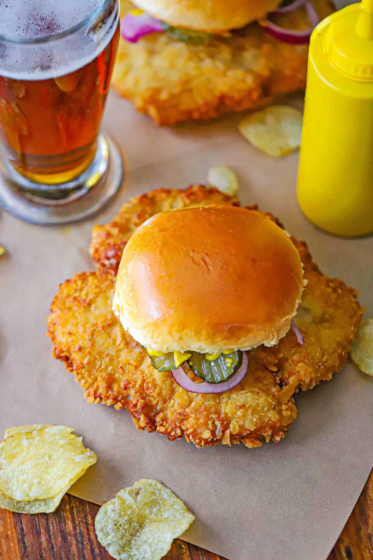 An overhead view of a breaded pork tenderloin sandwich including a crispy pork tenderloin red onion, pickles, yellow mustard, all on a hamburger bun with a bottle of yellow mustard and a glass of bear nearby.