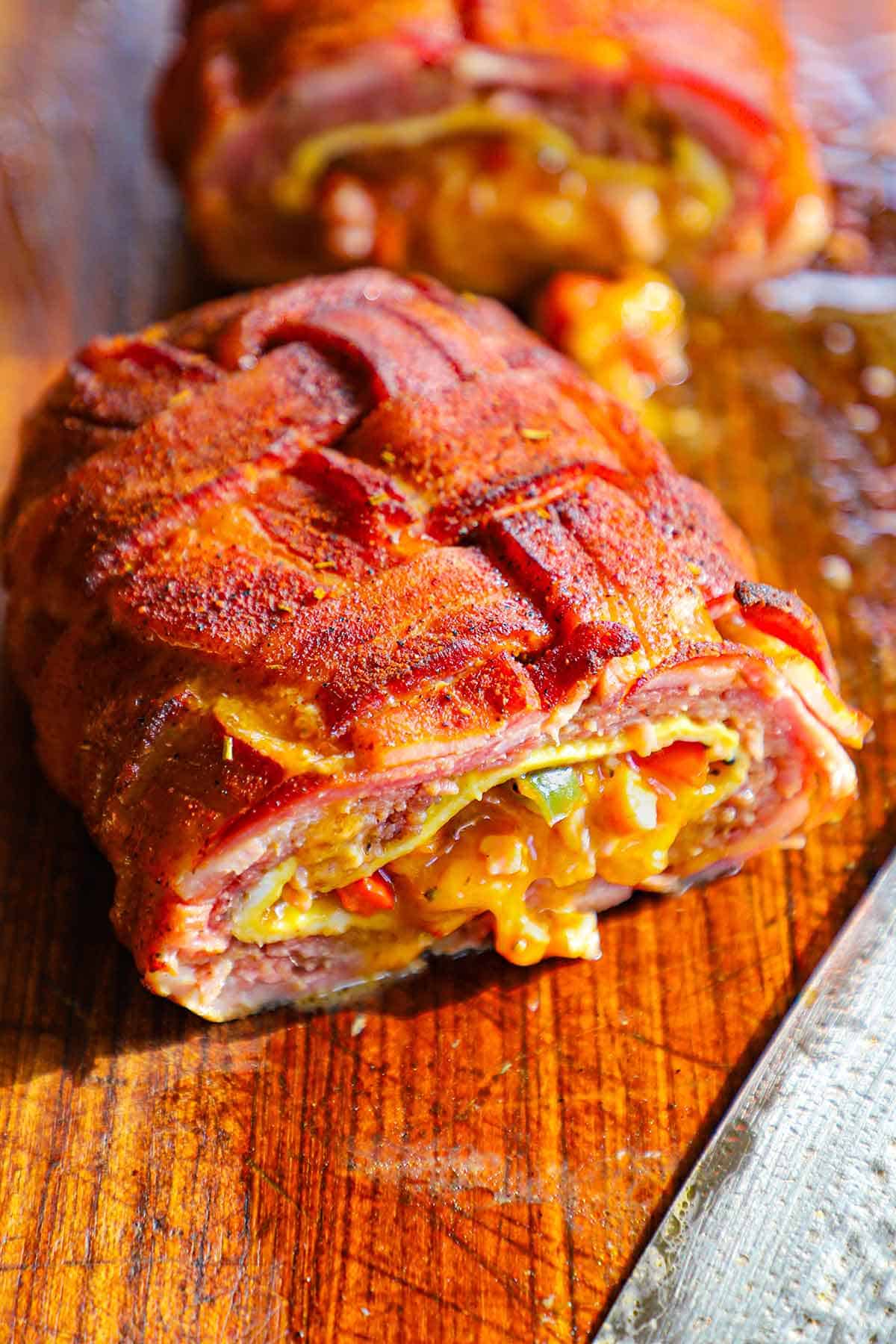 A close-up view of a smoked Western omelette fatty that has been cut in half revealing the melty cheddar cheese, saut&eacute;ed vegetables, cooked eggs, and juicy sausage, all wrapped in a seasoned bacon weave.
