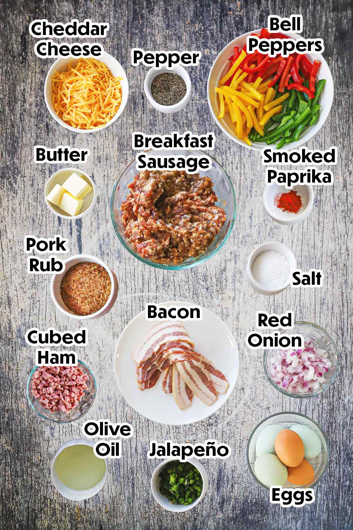 An arrangement of ingredients for a smoked Western Omelette Fatty on a grey wooden background including breakfast sausage, bell peppers, cheddar cheese, onion, cubed ham, eggs, jalapeño, bacon, and seasonings.