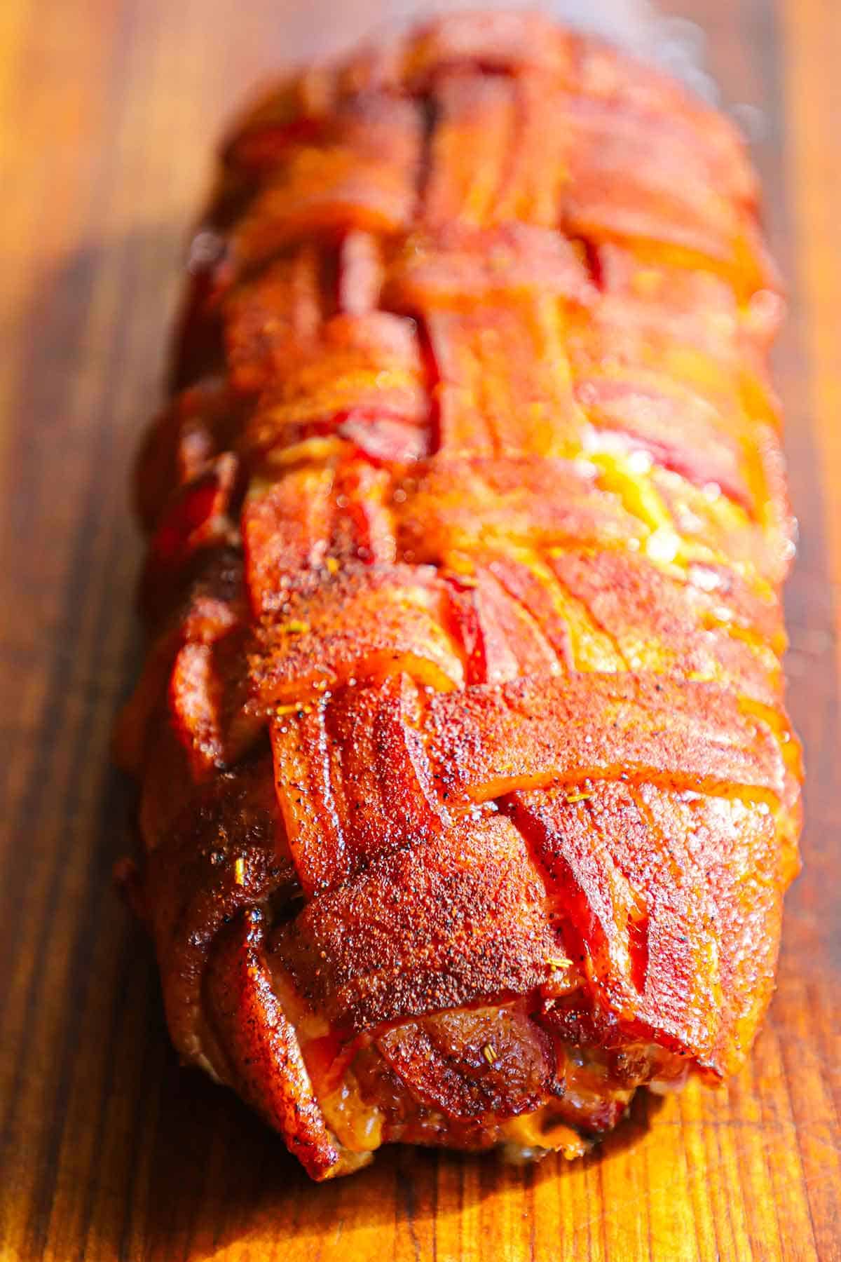 A straight-on view of a fully cooked smoked Western omelette Fatty that has been encased in a season bacon weave and is resting on a wooden cutting board.