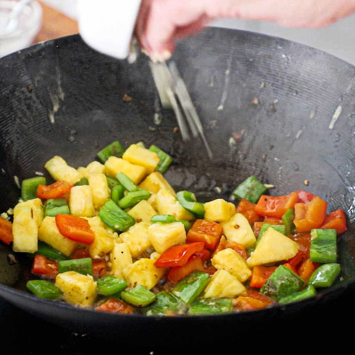 A person adding minced garlic and ginger into a wok that is filled with simmering red and green bell peppers and chunks to pineapple.