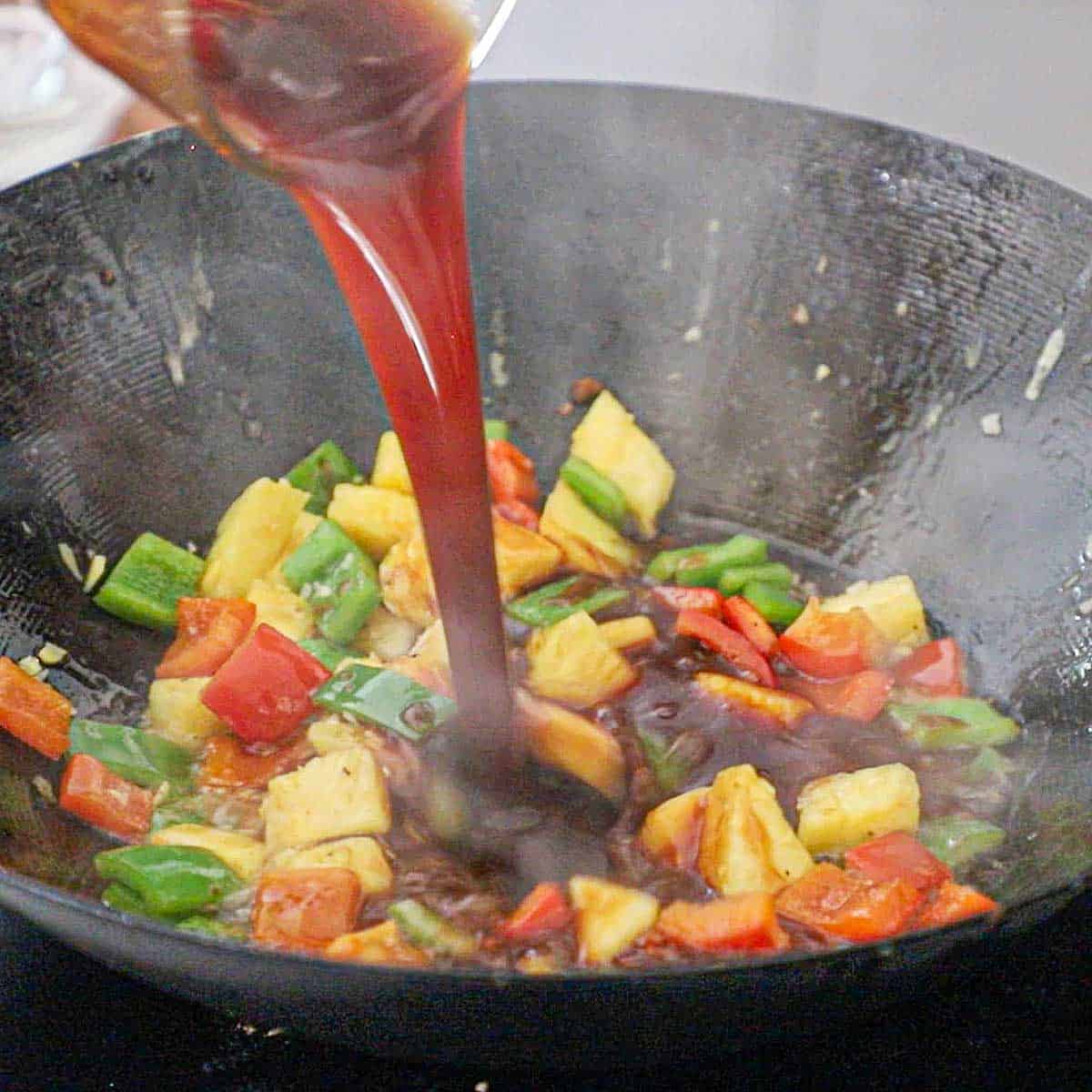 A person pouring a sweet and sour sauce from a small glass bowl into a wok filled with sautéed vegetables on pineapple.