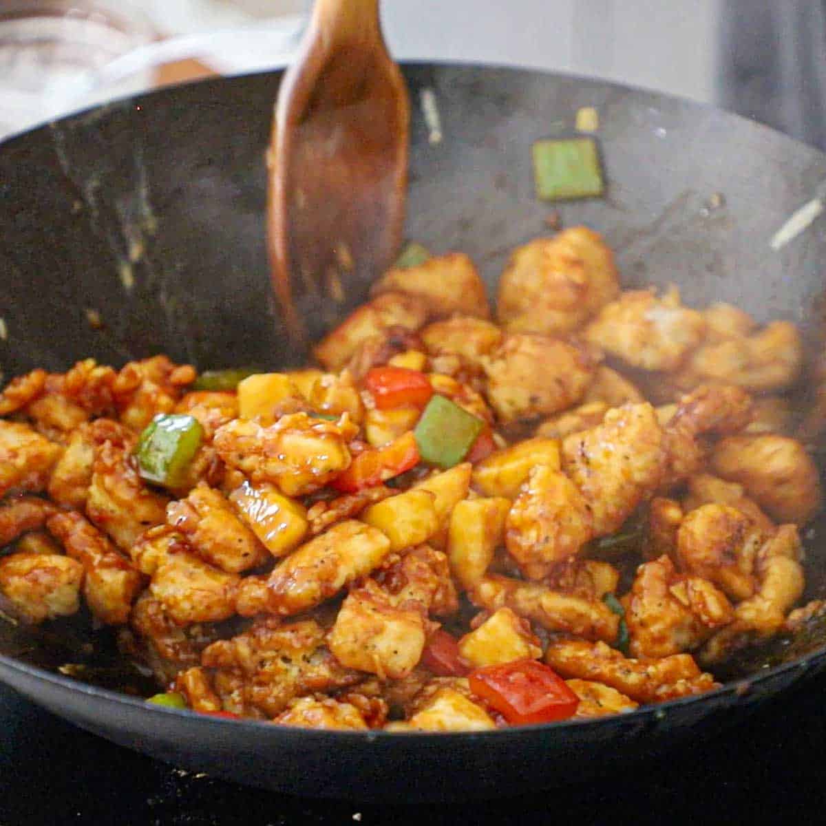 A person using two large wooden spoons to toss lightly fried piece of chicken in wok filled with sautéed vegetables and pineapple in a sweet and sour sauce.