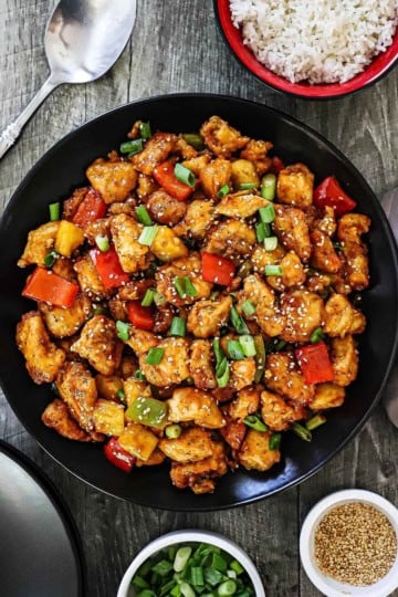An overhead view of a large black dinner plate filled with Classic Sweet and Sour Chicken Stir-Fry with a bowl of cooked white rice nearby.