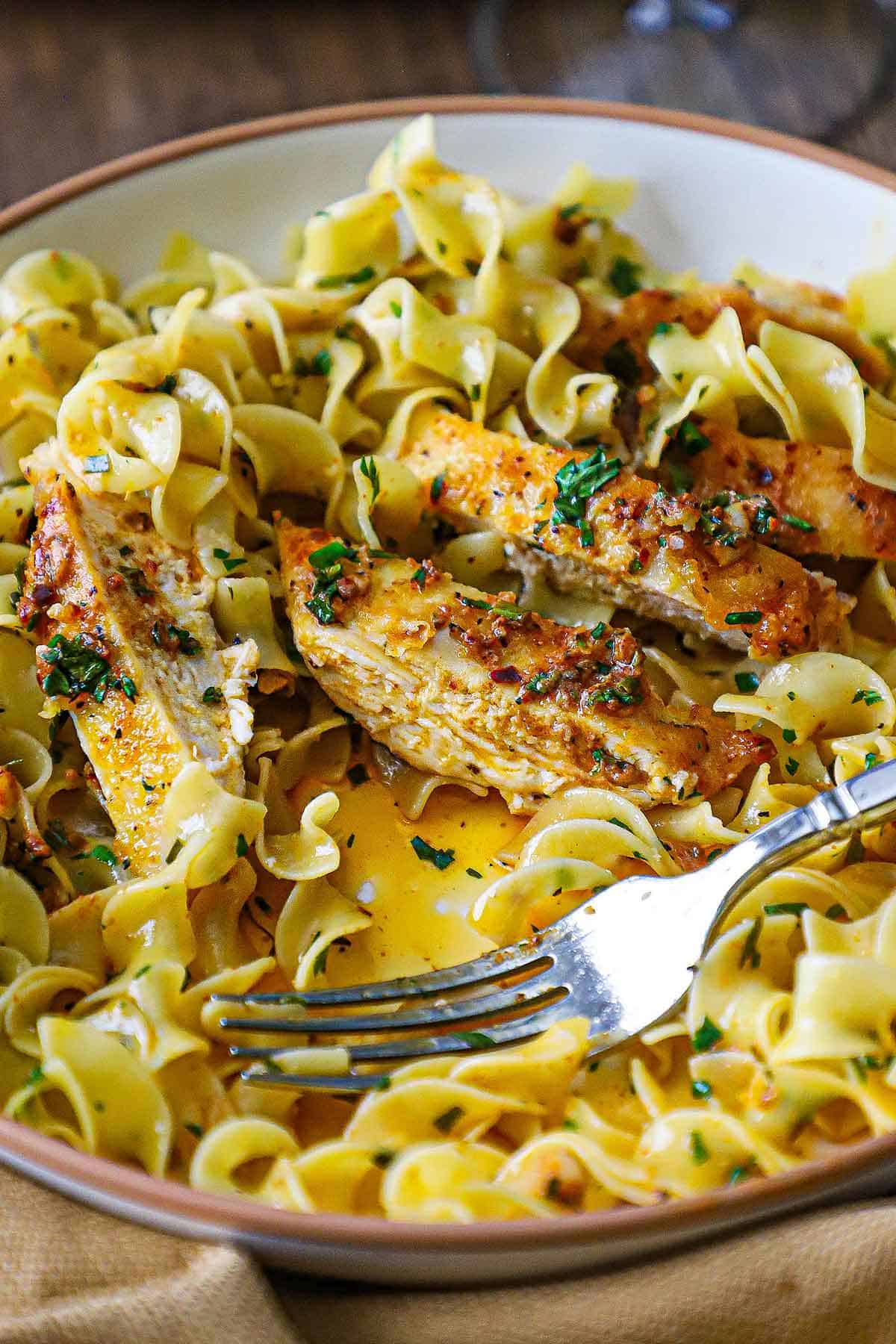 A close-up view of a shallow pasta bowl filled with a partially eaten serving of Cowboy Butter Chicken with egg noodles and with a fork resting in the bowl.