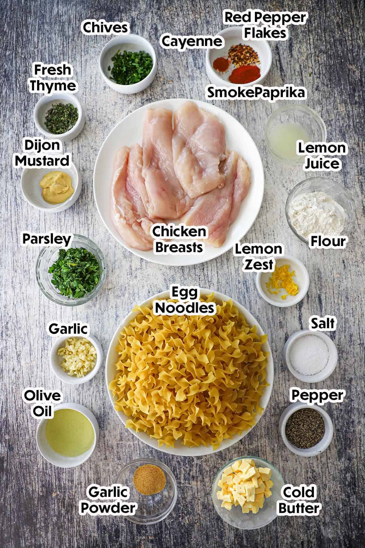 An arrangement of ingredients for Cowboy Butter Chicken on a grey wooden background including chicken breasts, egg noodles, cold butter, garlic, flour, olive oil, herbs, and spices.