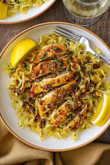 An overhead view of a shallow pasta bowl filled with a serving of Cowboy Butter Chicken with Egg Noodles with two lemon wedges on each side of the dish and a glass of white wine nearby.