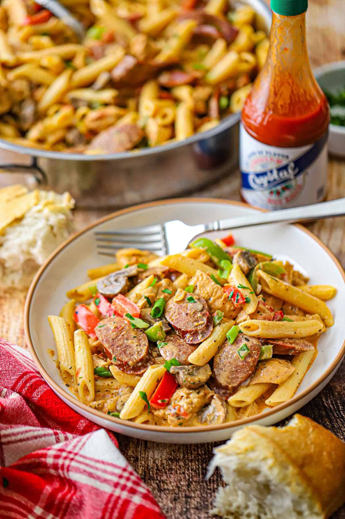 A straight-on view of a shallow pasta bowl filled with a serving of Creamy Cajun Chicken and Sausage Pasta in it with a bottle of hot sauce nearby.