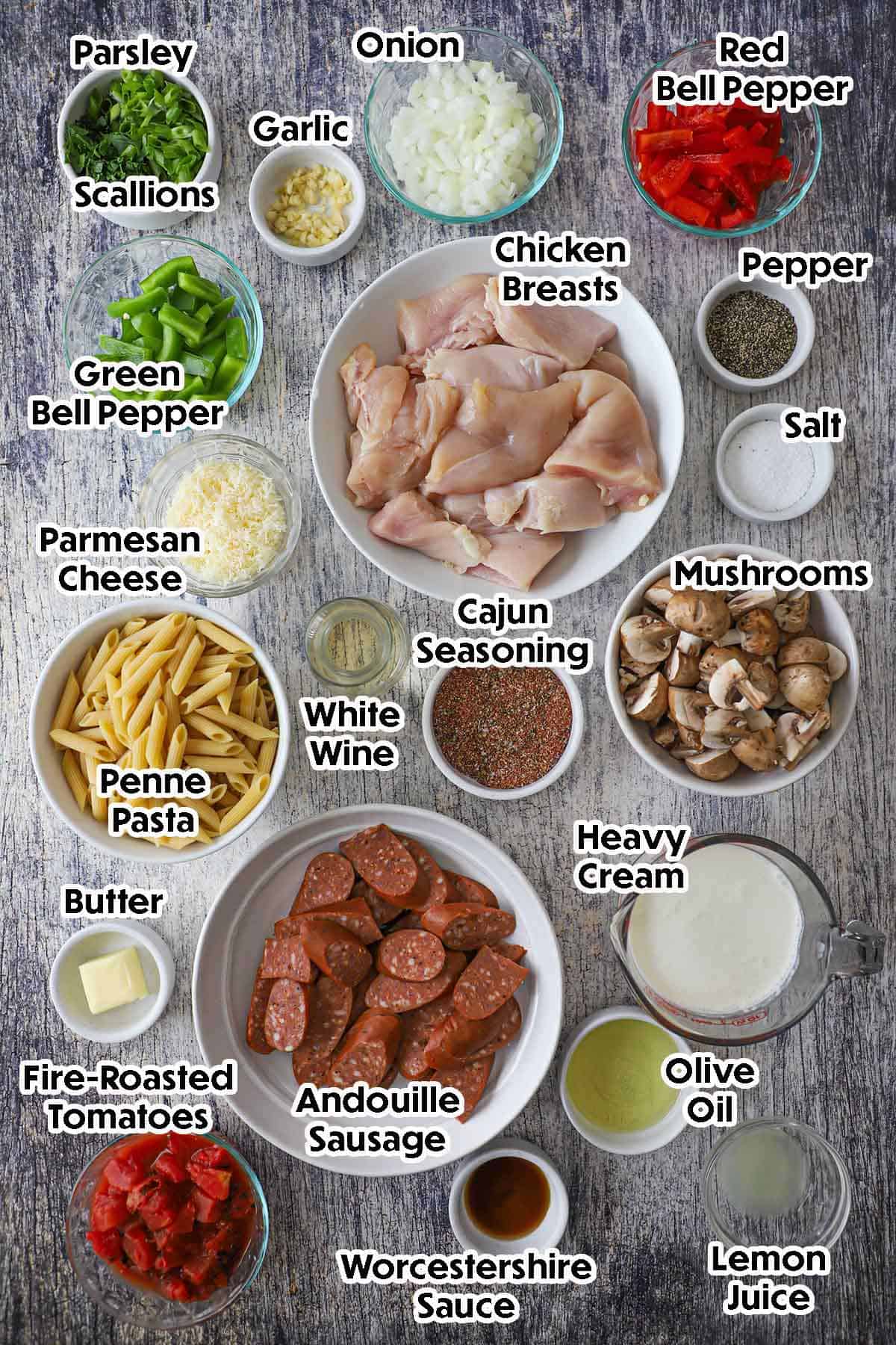 An arrangement of ingredients for Creamy Cajun Chicken and Sausage Pasta on a grey-wooden background including chicken breasts, sausage, mushrooms, bell peppers, onion, garlic, pasta, cream, tomatoes, wine, herbs, and seasonings.