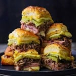 A close-up view of tall stack of Melty Cuban Sliders consisting of sliced ham, shredded pork, pickles, melty Swiss cheese, and yellow mustard on seasoned Hawaiian slider buns.