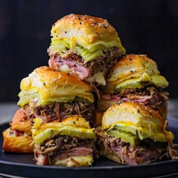 A close-up view of tall stack of Melty Cuban Sliders consisting of sliced ham, shredded pork, pickles, melty Swiss cheese, and yellow mustard on seasoned Hawaiian slider buns.