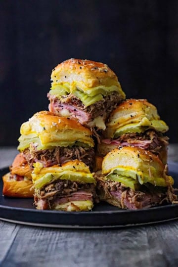 A close-up view of tall stack of Melty Cuban Sliders consisting of sliced ham, shredded pork, pickles, melty Swiss cheese, and yellow mustard on seasoned Hawaiian slider buns.