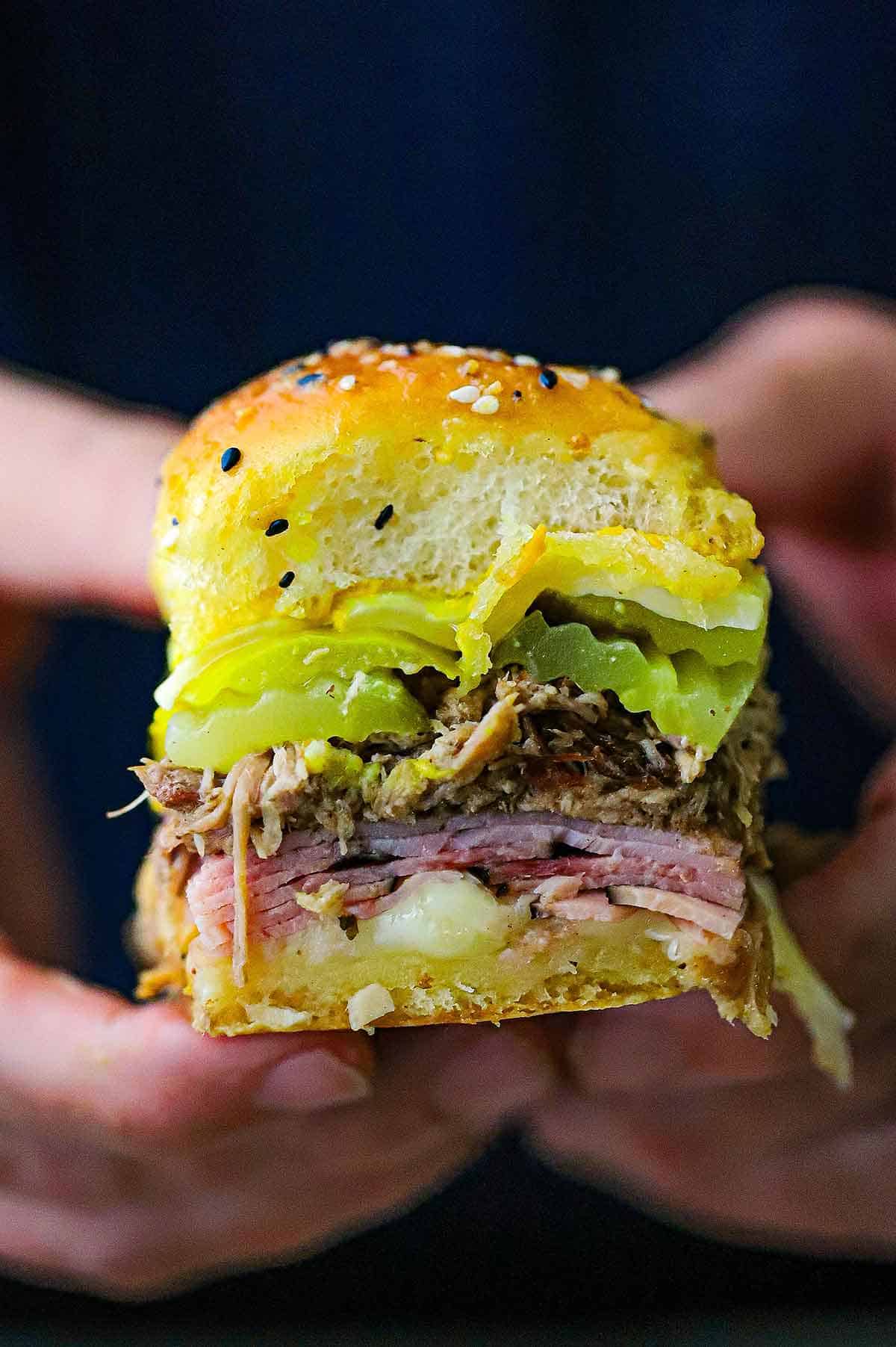 A person using both hands to hold a Cuban Slider in his hand consisting of sliced ham, shredded pork, pickles, melty Swiss cheese, and yellow mustard on a seasoned Hawaiian slider bun.