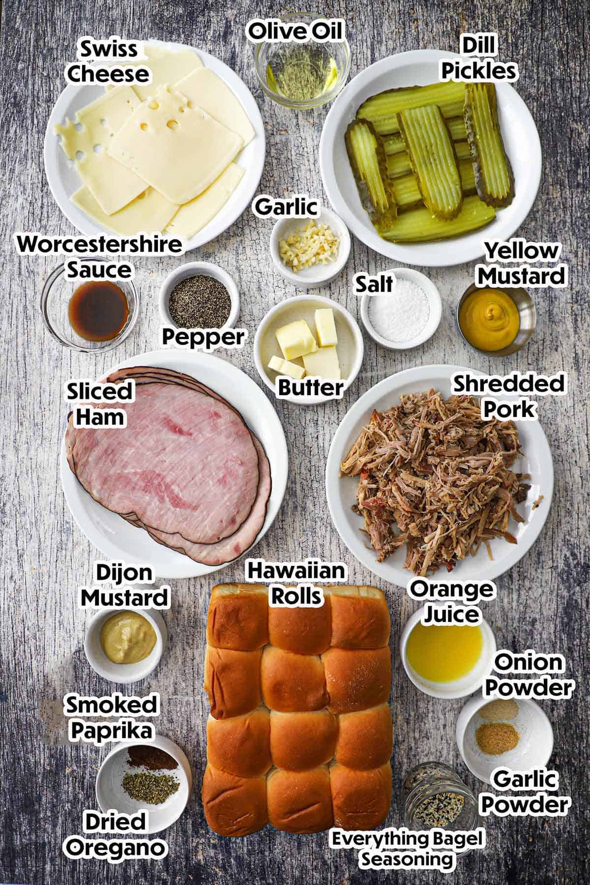 An arrangement of ingredients for Melty Cuban Sliders on a grey wooden background including sliced ham, shredded pork, dill pickles, garlic, Swiss cheese, butter, orange juice, mustard, Hawaiian rolls, and seasonings.