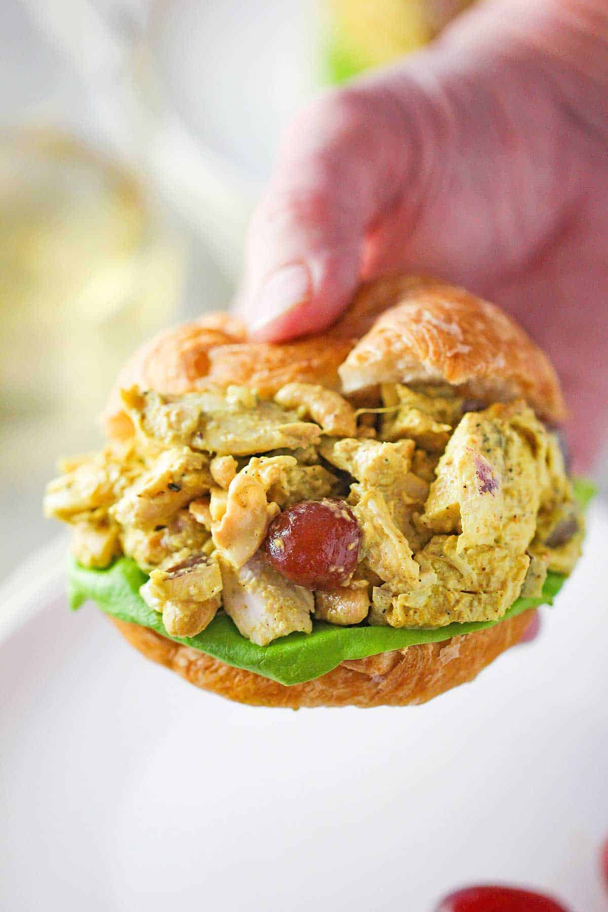 A person holding a croissant filled with Curried Chicken Salad and a leaf of green lettuce.