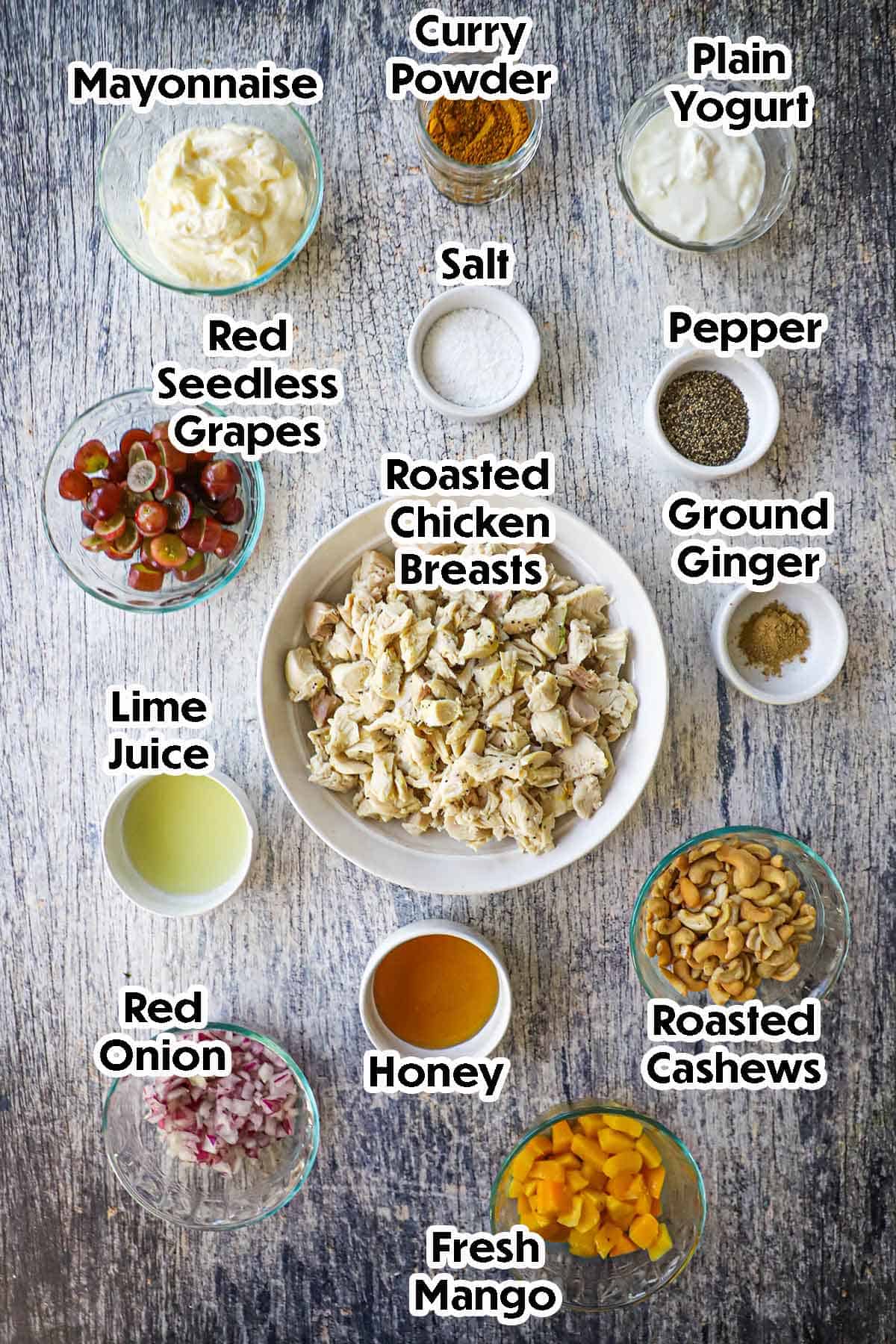 An arrangement of ingredients for Curried Chicken Salad on a grey wooden background including chopped roasted chicken, grapes, mayonnaise, yogurt, cashews, red onion, mango, lime juice, and spices and seasonings.