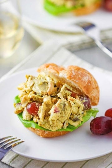 A straight-on view of a dinner roll split open and filled with Curried Chicken Salad on a leaf of green lettuce on a white plate with a couple of red grapes resting nearby.