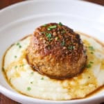 A straight-on view of a large Dutch meatball resting on a bed of mashed potatoes with a thin brown gravy drizzled over the top all in a white shallow bowl.