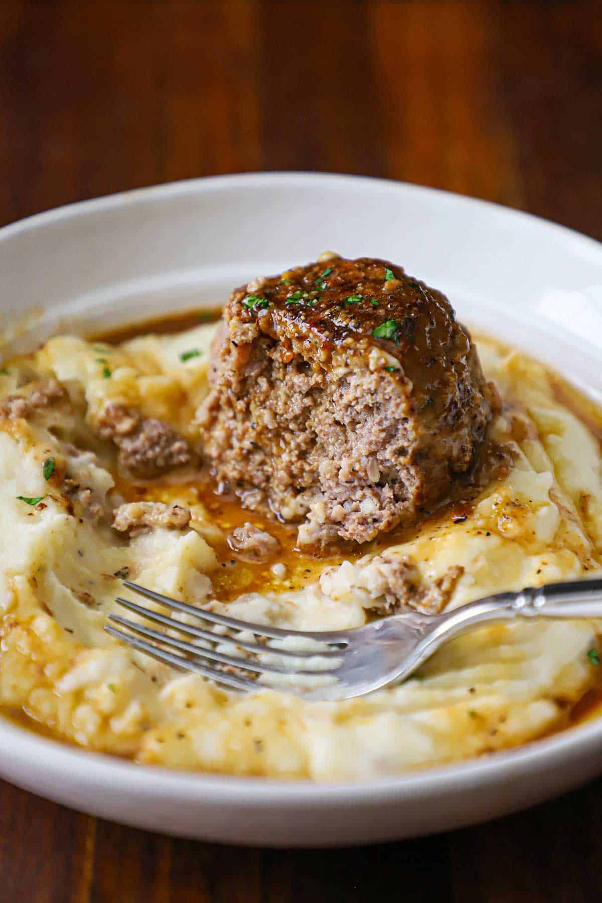 A half eaten Dutch meatball that is topped with a brown gravy and is resting on a partially eaten bed of mashed potatoes in a shallow white bowl.