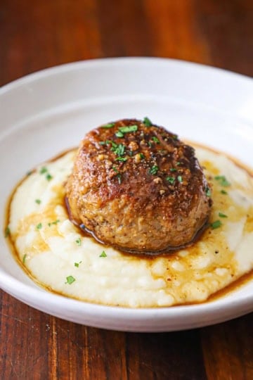 A straight-on view of a large Dutch meatball resting on a bed of mashed potatoes with a thin brown gravy drizzled over the top all in a white shallow bowl.