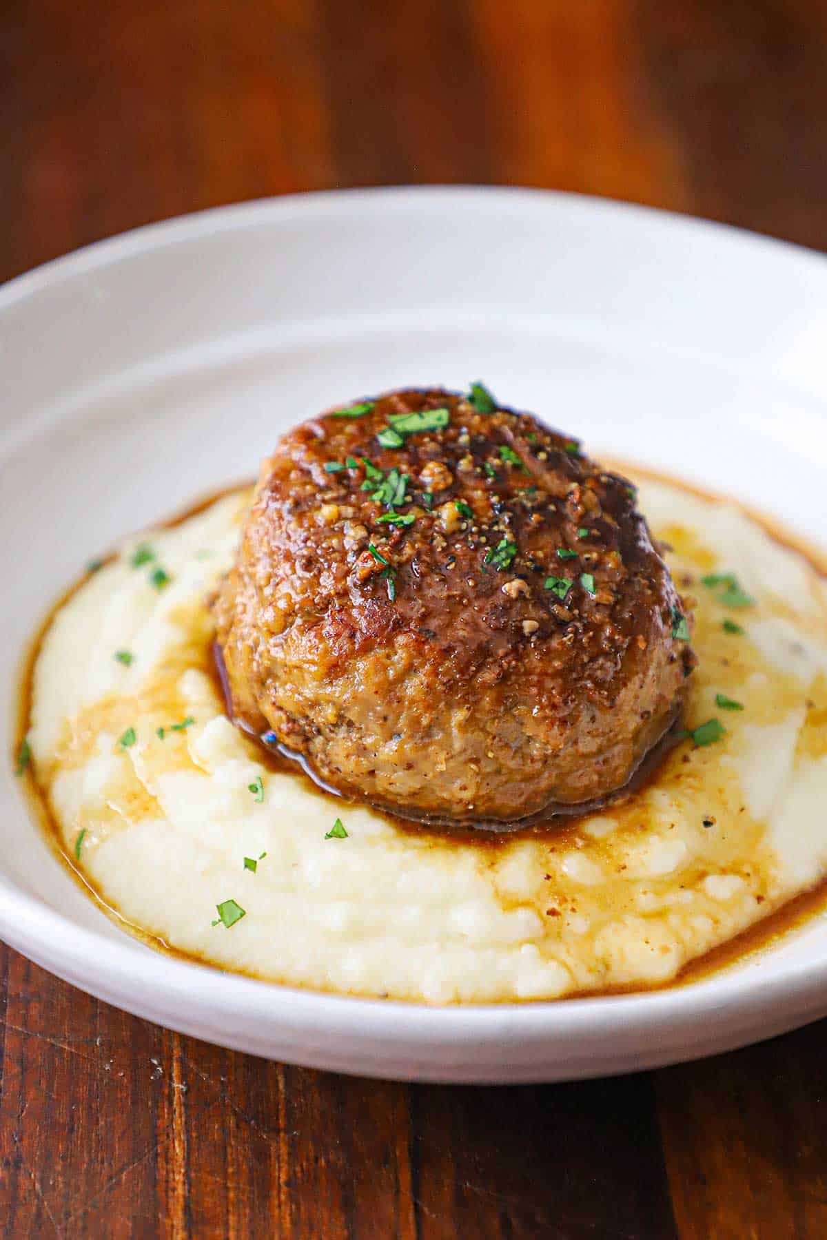 A straight-on view of a large Dutch meatball resting on a bed of mashed potatoes with a thin brown gravy drizzled over the top all in a white shallow bowl.