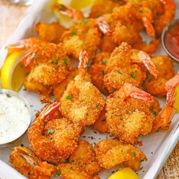 An overhead view of Fried Butterfly Shrimp on a metal pan lined with white wax paper with a small bowl of cocktail sauce in one corner and and a small bowl of tartar sauce in the other corner.