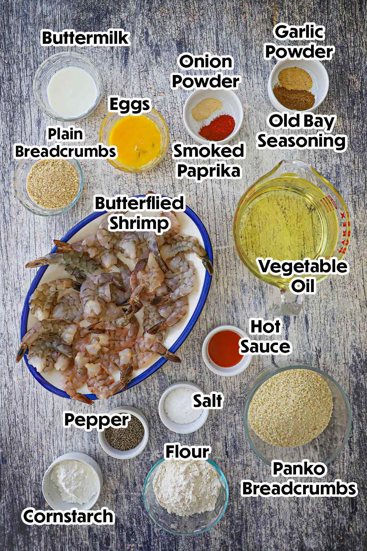 An arrangement of ingredients for Fried Butterfly Shrimp on a grey wooden background including butterflied raw shrimp, flour, cornstarch, eggs, buttermilk, breadcrumbs, oil, and seasonings.