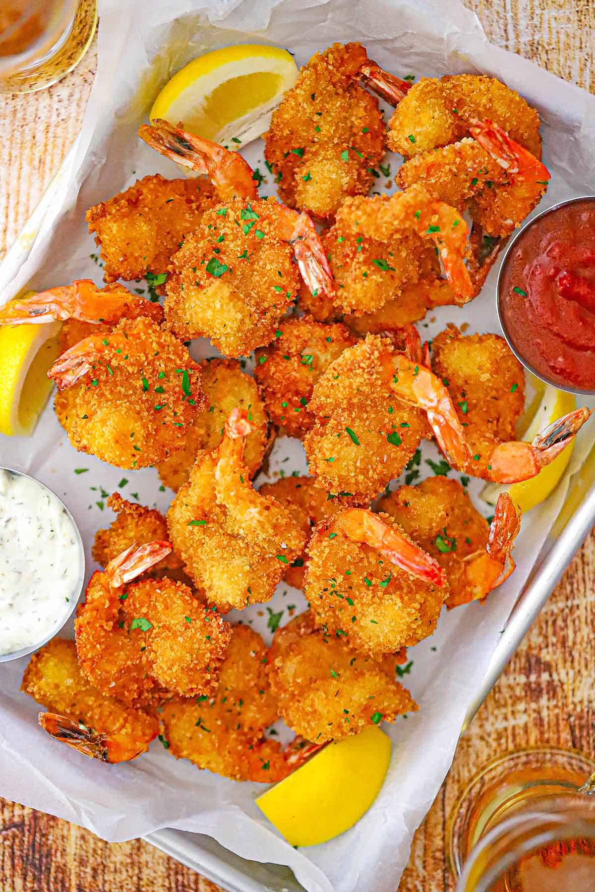 An overhead view of Fried Butterfly Shrimp on a metal pan lined with white wax paper with a small bowl of cocktail sauce in one corner and and a small bowl of tartar sauce in the other corner.