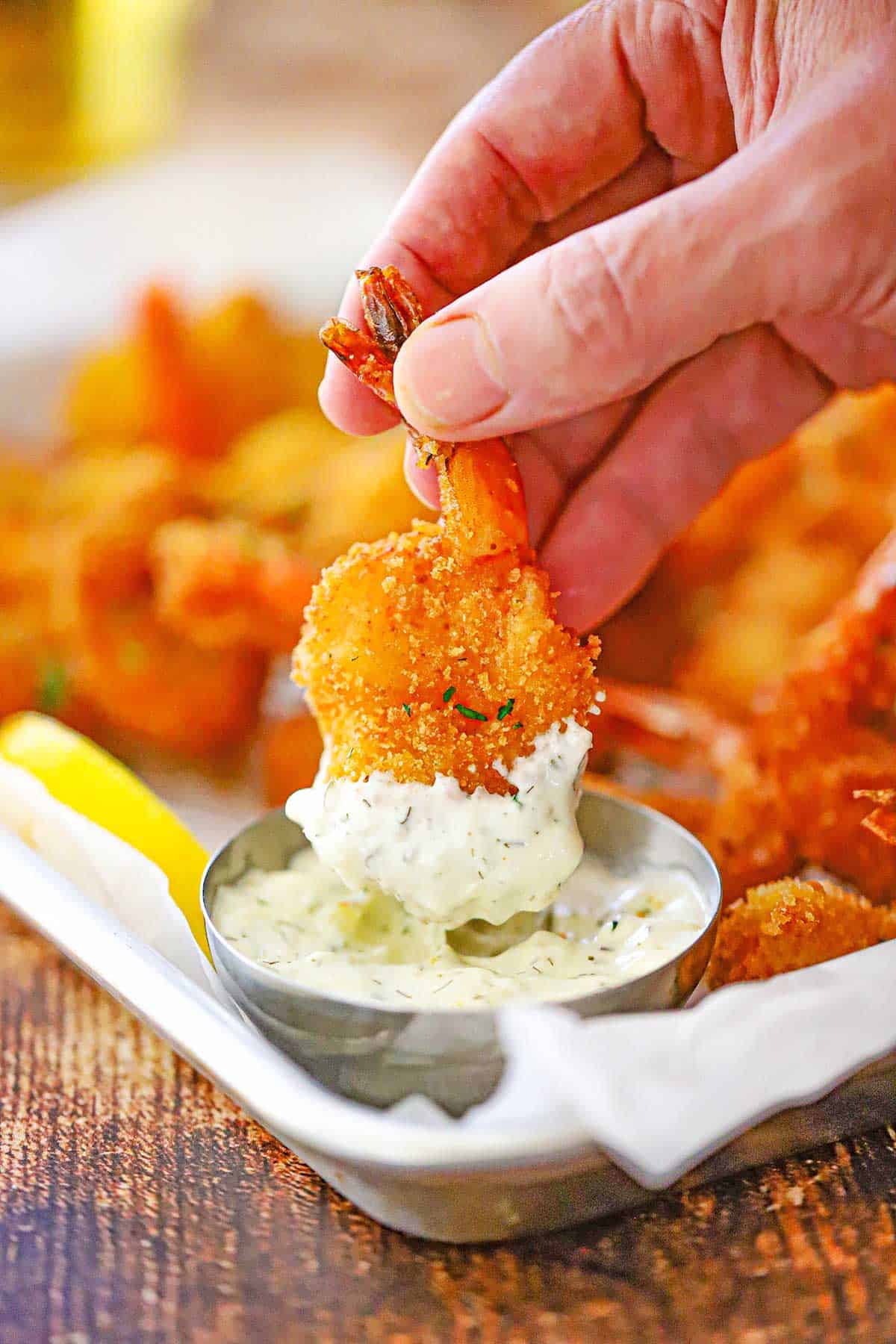A person using his fingers to plunge a single Fried Butterfly Shrimp into a small silver bowl filled with homemade tartar sauce.