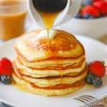 A close-up view of a stack of Fluffy Buttermilk Pancakes on a white plate with fresh fruit next to the pancakes and maple syrup being poured over the top.