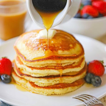 A close-up view of a stack of Fluffy Buttermilk Pancakes on a white plate with fresh fruit next to the pancakes and maple syrup being poured over the top.
