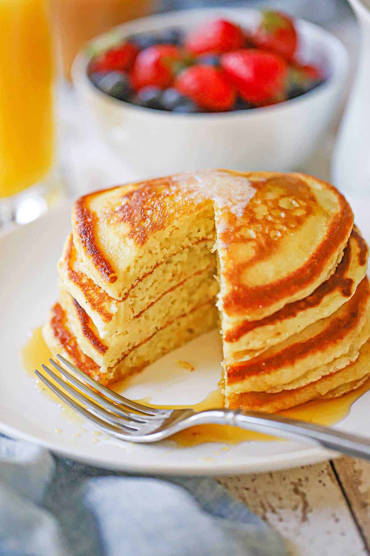 A stack of four fluffy buttermilk pancakes on a white plate sitting in a small pool of maple syrup and there is a triangular pice of the pancakes missing from the stack.