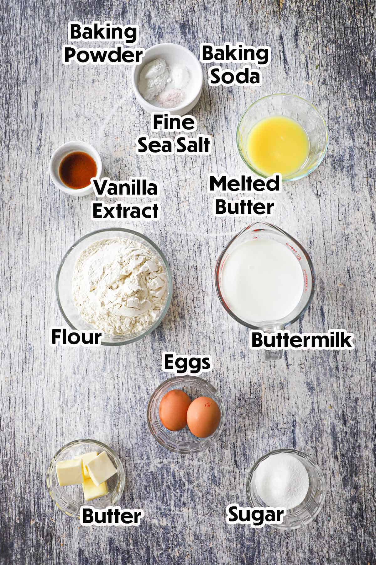 An arrangement of ingredients for Fluffy Buttermilk Pancakes on a grey wooden background including flour, baking powder, baking soda, salt, buttermilk, butter, eggs, and sugar.