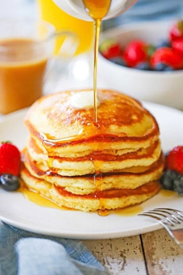 A straight-on view of a stack of Fluffy Buttermilk Pancakes on a white plate with fresh fruit next to the pancakes and maple syrup being poured over the top.