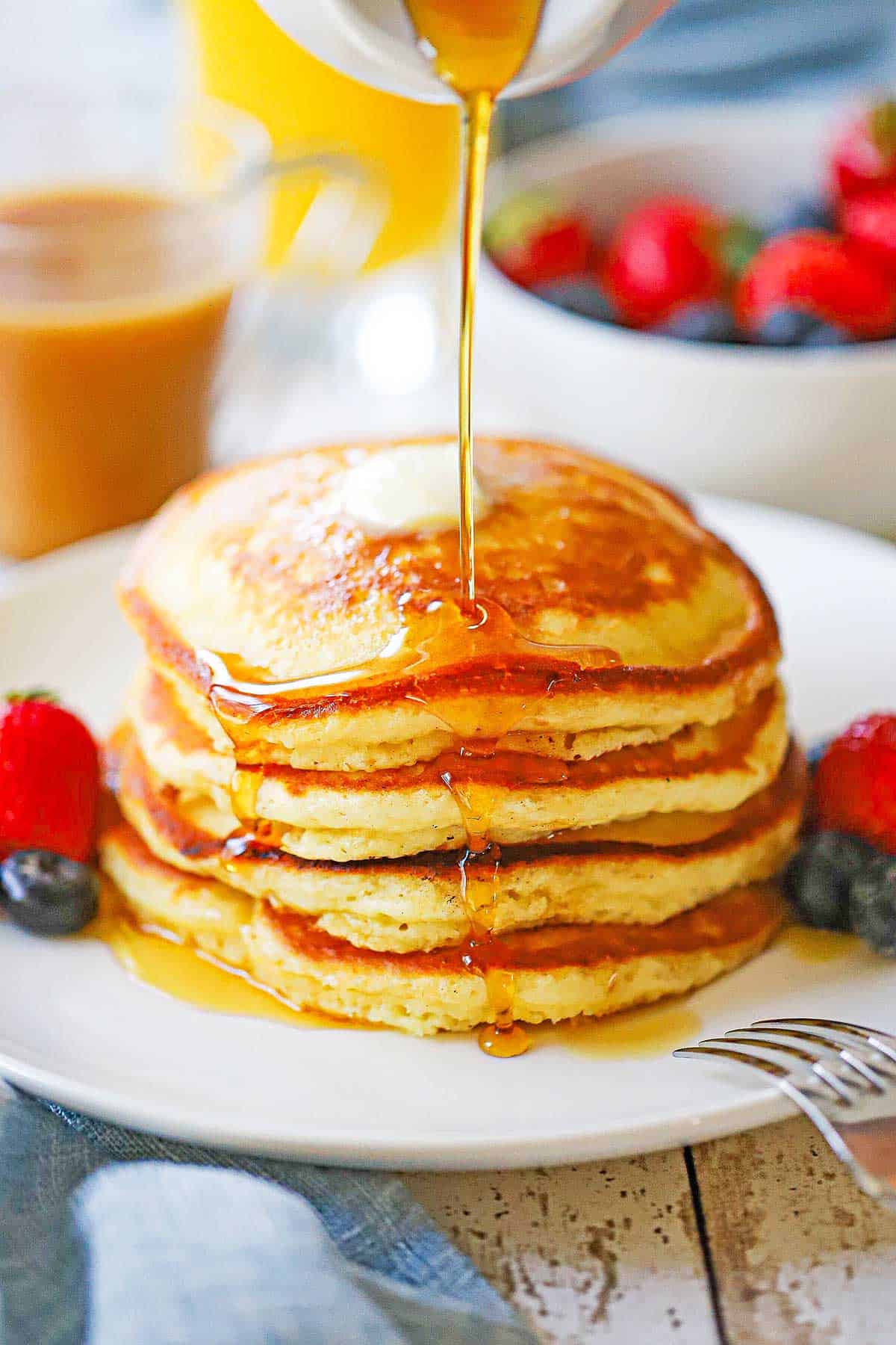 A straight-on view of a stack of Fluffy Buttermilk Pancakes on a white plate with fresh fruit next to the pancakes and maple syrup being poured over the top.