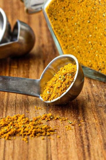 A small metal teaspoon filled with homemade curry powder on a wooden cutting board with more of the curry powder in a container nearby.