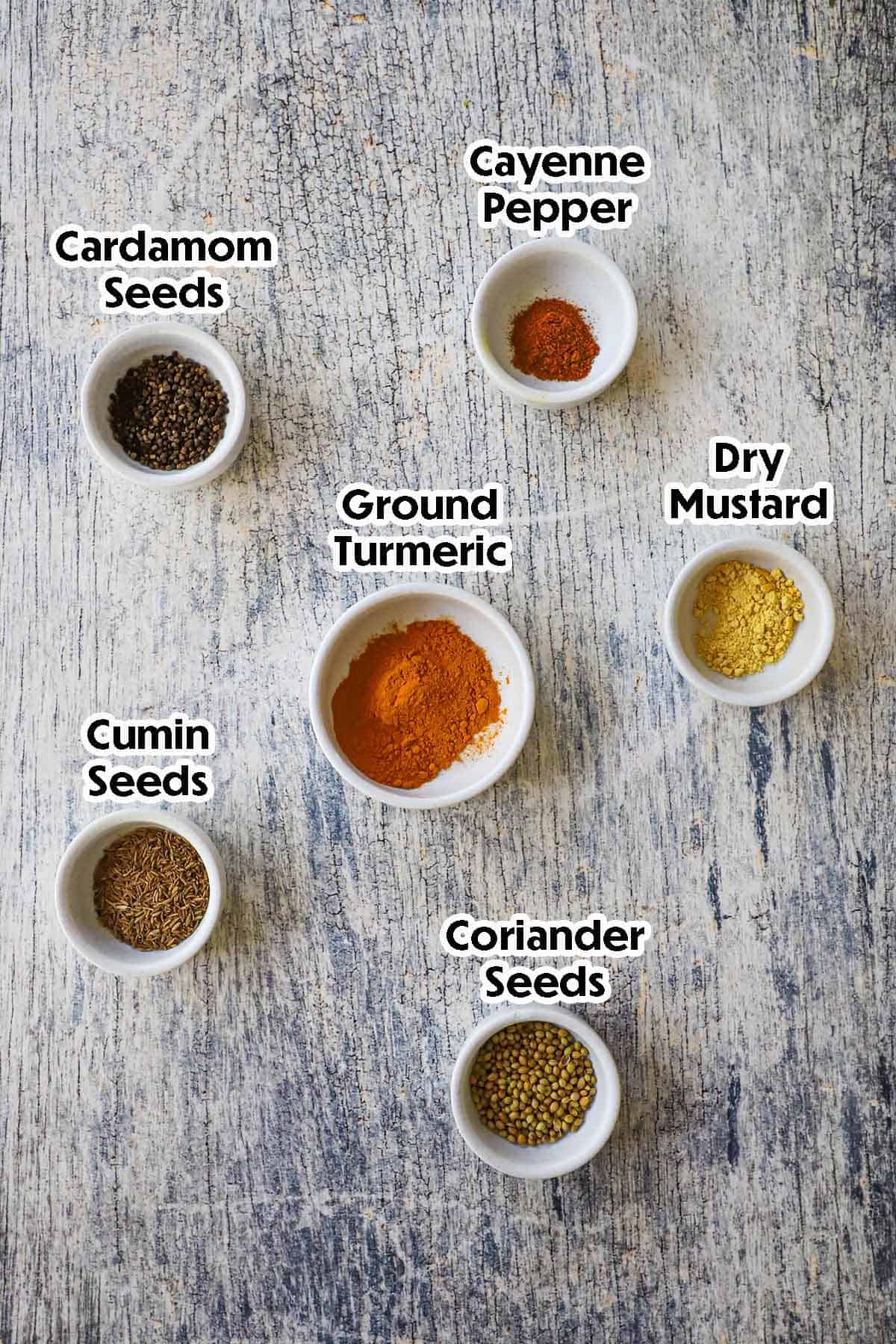 An arrangement of ingredients for homemade curry powder on a grey wooden background including ground turmeric, dry mustard, cayenne pepper, cardamom seeds, cumin seeds, and coriander seeds.