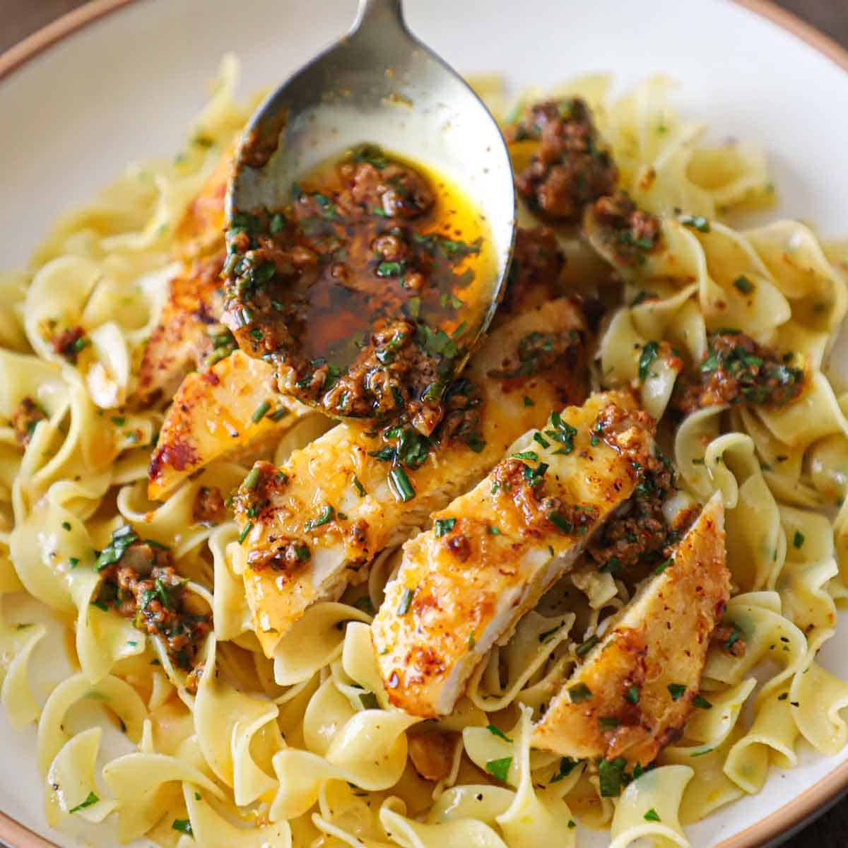 A person using a serving spoon to drizzle an herbed Cowboy butter sauce of slices of seared chicken breast resting on buttered noodles in a shallow pasta dish.